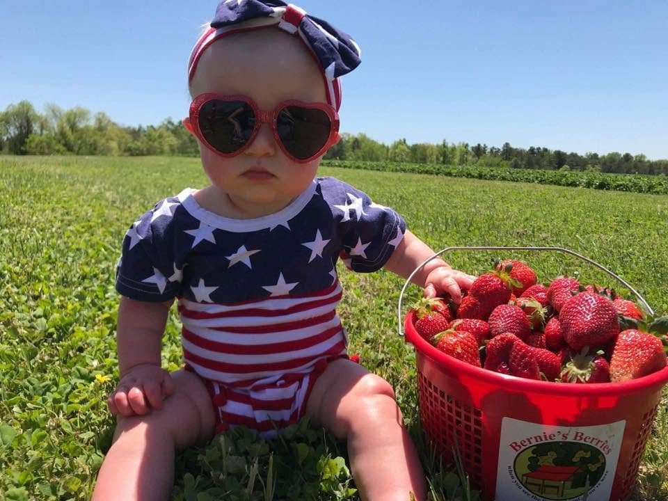 A baby in heart sunglasses sitting in the field next to a basket of strawberries