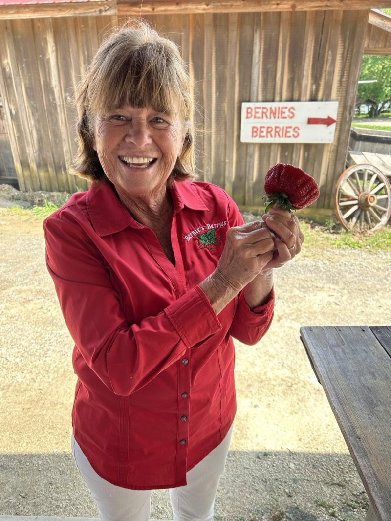 Bernie smiling outside the barn, holding up a ripe strawberry